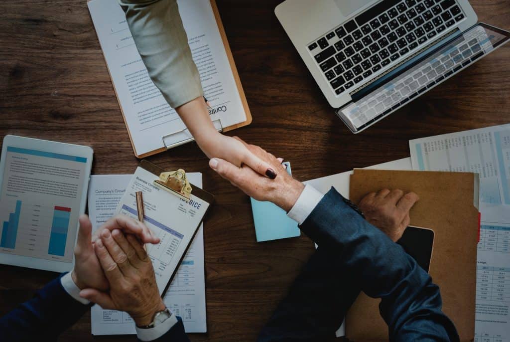 Two people shake hands over a wooden desk with documents, a laptop, and a tablet, indicating a business agreement.