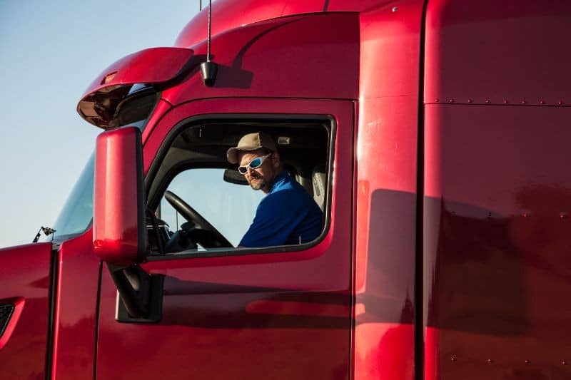 A man wearing sunglasses and a hat is sitting in the driver's seat of a red semi-truck, looking out the open window.