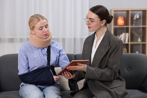 A woman with a neck brace and arm sling sits on a couch while another woman in a suit shows her something on a tablet.