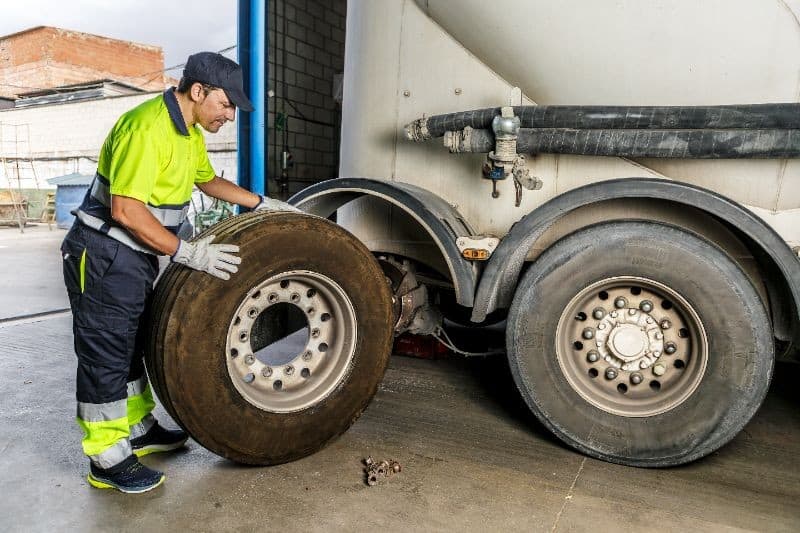 A worker in safety gear prepares to install a large truck tire next to a parked truck, with tools on the ground nearby.