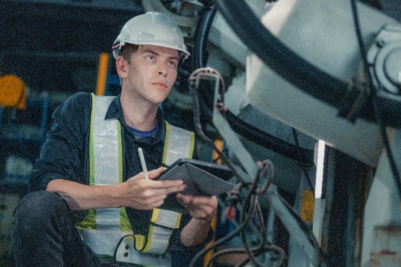 A worker wearing a safety helmet and reflective vest inspects machinery while holding a tablet and stylus in an industrial setting.