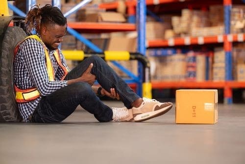 A warehouse worker in a safety vest sits on the floor, holding his leg in pain next to a cardboard box, with shelves in the background.