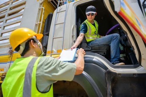 A worker in a safety vest and helmet hands a clipboard to a truck driver seated in the cab of a large vehicle.