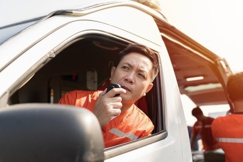 A person in an orange uniform sits in a vehicle, speaking into a handheld radio, with another person visible outside near the vehicle.