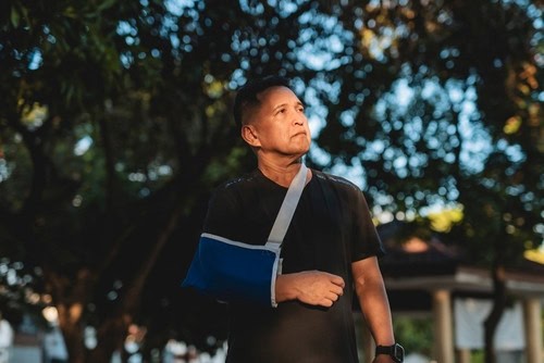A man with a blue arm sling stands outdoors in a park, looking off into the distance in the late afternoon light.