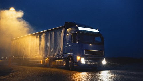 A large semi-truck with headlights on drives on a wet road at night under a streetlight, with mist in the air.