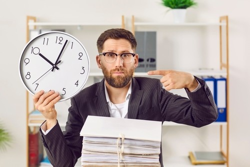A man in a suit holding a large clock and pointing at it, with a stack of paperwork in front of him in an office setting.