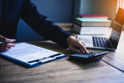 Person in business attire using a calculator next to a clipboard with documents, a pen, a laptop, and stacked books on a wooden desk.