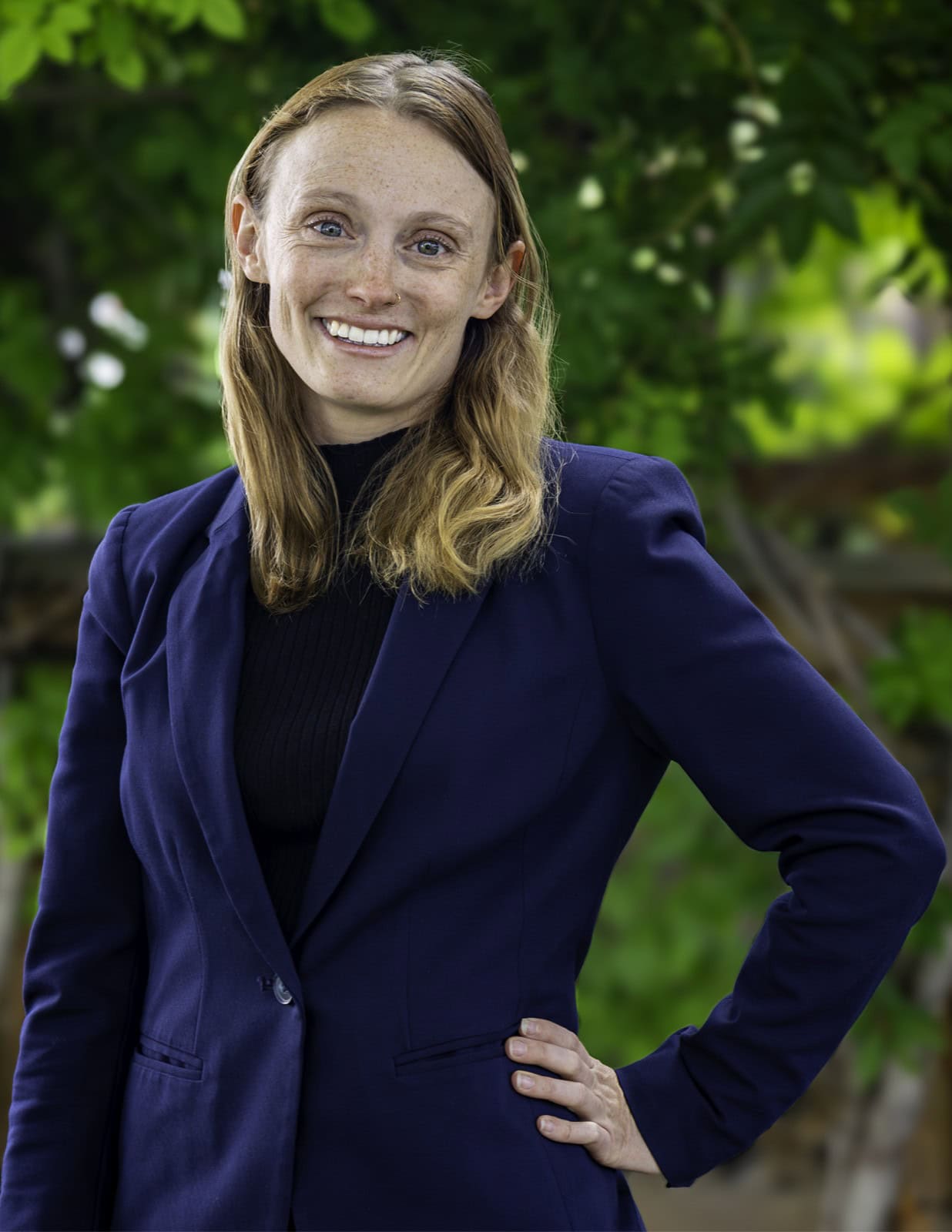 A woman with light brown hair wearing a navy blazer and black top stands outdoors in front of green foliage, smiling at the camera with one hand on her hip.