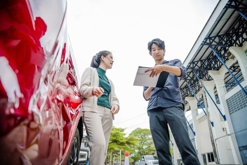 A man holding a clipboard talks with a woman next to a red car with a dented door, outdoors by a building.