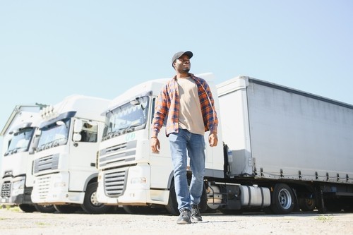A man in casual clothes and a cap walks in front of a row of parked white semi-trailer trucks on a sunny day.