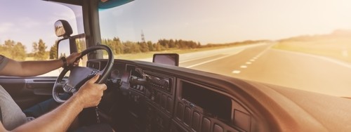 Person driving a truck on an open, empty highway in daylight, viewed from inside the cab with hands on the steering wheel.