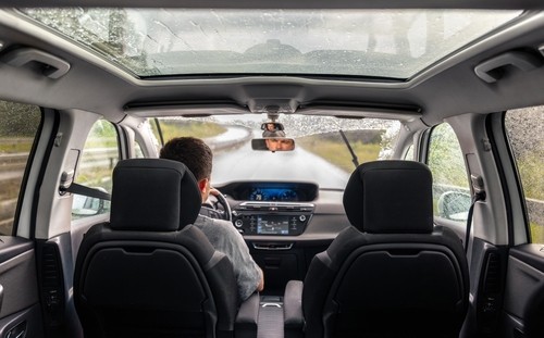 A person drives a car on a rainy day, viewed from the back seat. The windshield wipers are active and the sunroof shows raindrops.