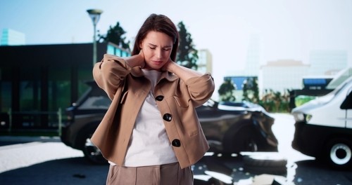 A woman stands holding her neck in apparent pain or discomfort with two damaged vehicles behind her, suggesting a recent car accident.
