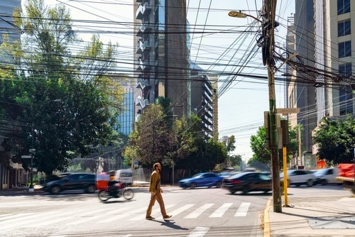 A person crosses a city street at a crosswalk while cars and a motorcycle pass by, with numerous overhead power lines and tall buildings in the background.