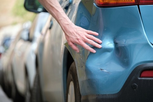 A hand points to a dented and scratched area on the rear side of a blue car parked on the street.